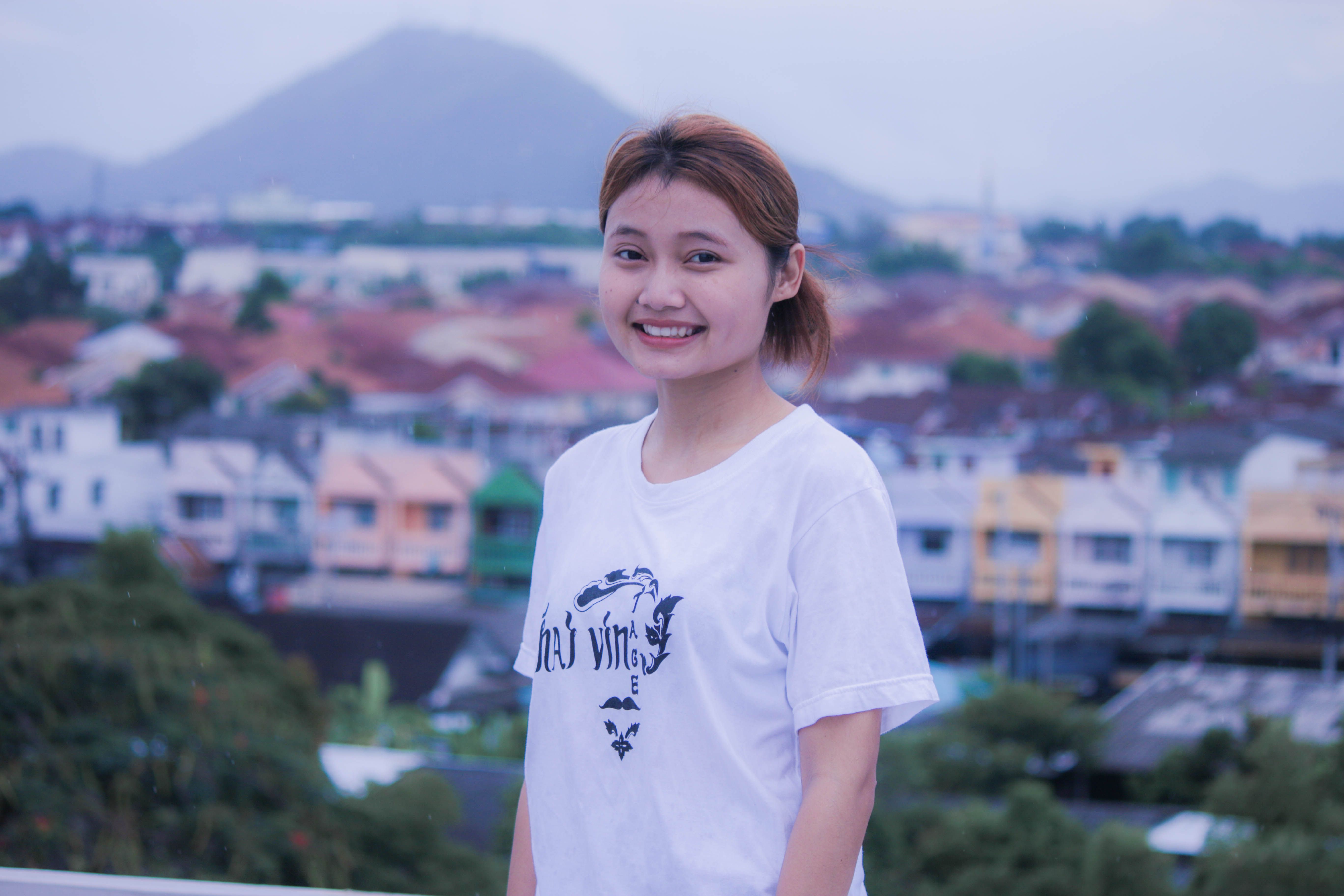 Smiling young woman in a white shirt with a blurred cityscape and mountains in the background.