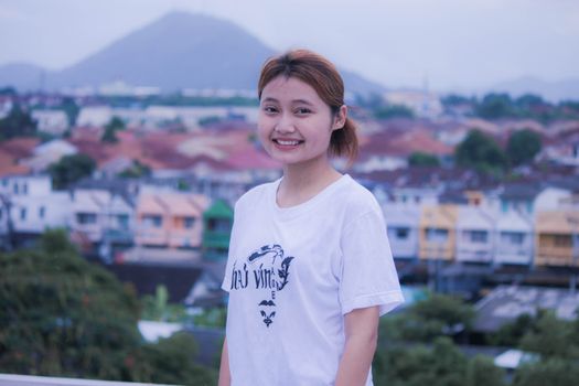 Smiling young woman in a white shirt with a blurred cityscape and mountains in the background. - Fotografer Sidoarjo
