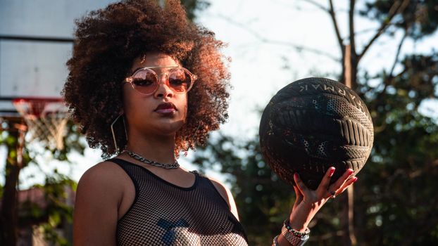 Woman with afro hair holding basketball in sunlight, wearing sunglasses.