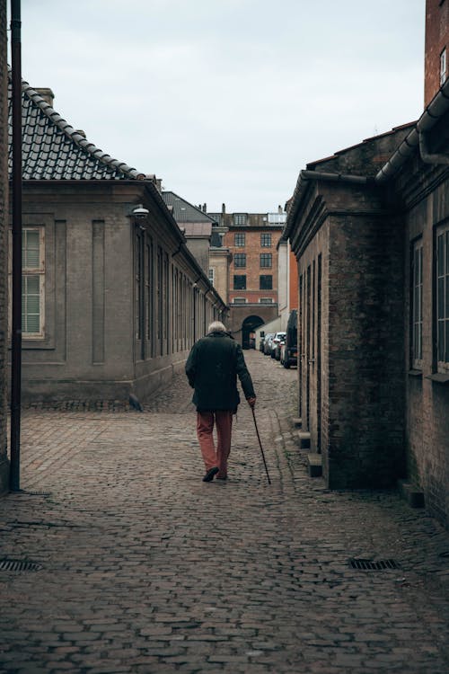 Back View of an Elderly Man Walking with a Stick Through a Cobblestone Street