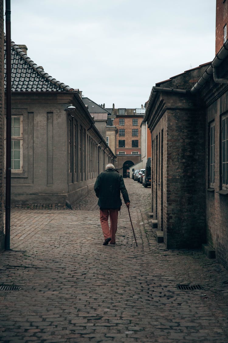 Back View Of An Elderly Man Walking With A Stick Through A Cobblestone Street