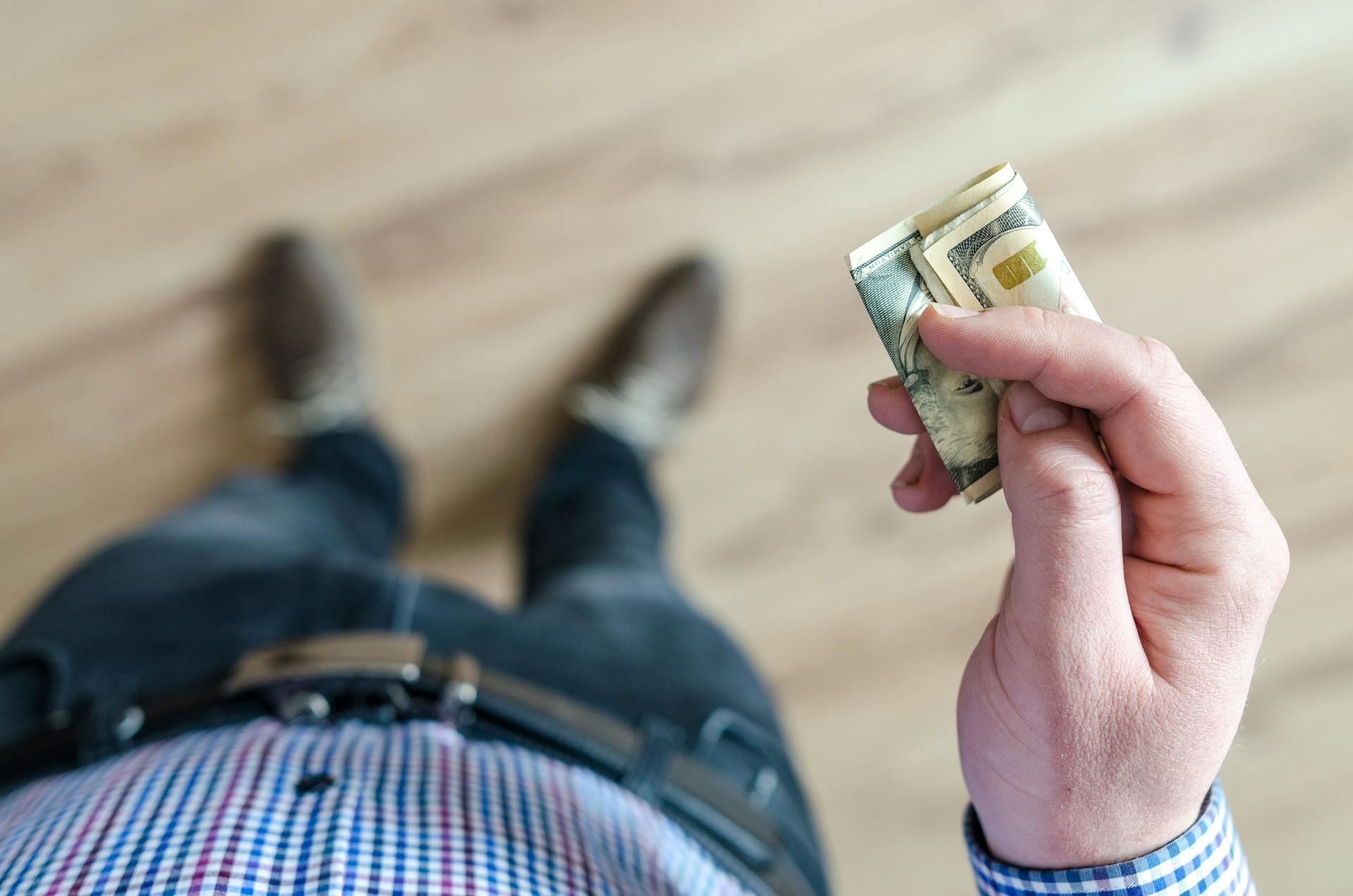 A person wearing denim and a checked shirt holds folded dollar bills in their hand indoors.