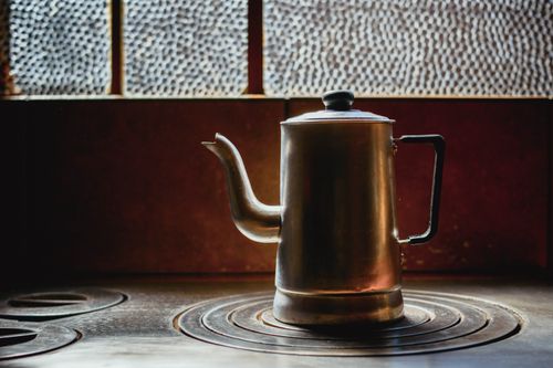 Free A solitary silver kettle on a rustic stove, bathed in warm light with a textured window backdrop. Stock Photo