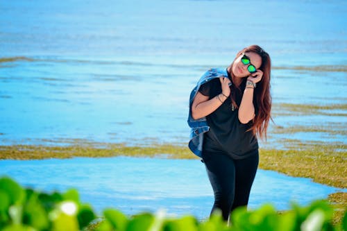 A Woman in Blue Denim Jacket and Black Pants Wearing Sunglasses Standing Beside Sea