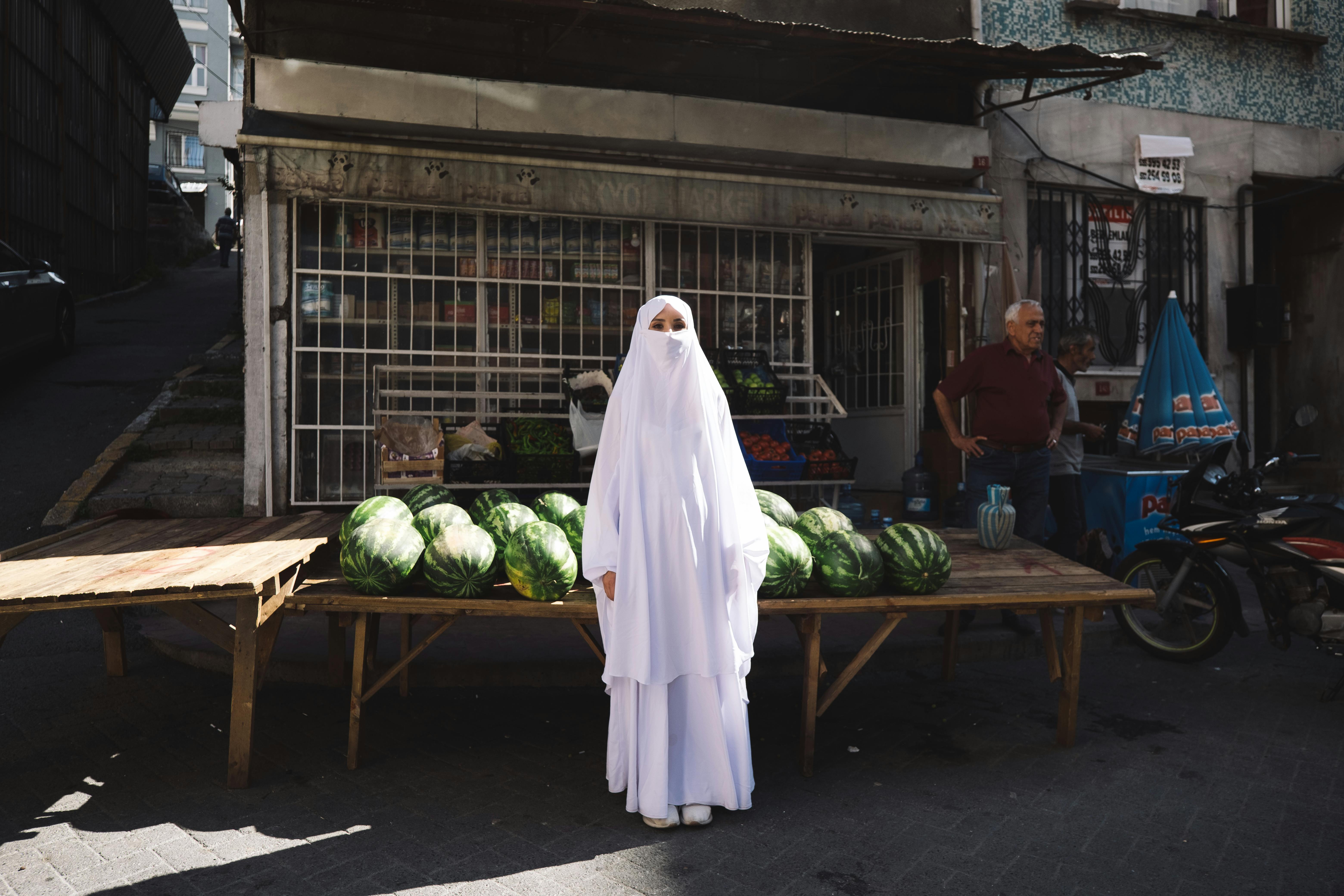 Woman in traditional attire stands with watermelons at a street market.