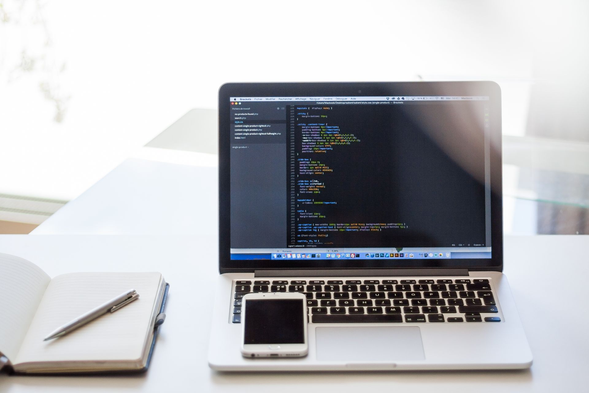 Open laptop displaying code on a desk with smartphone and notebook, ideal for technology and business use.