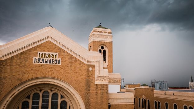 Brick facade of First Baptist Church with tower viewed against a dramatic cloudy sky.
