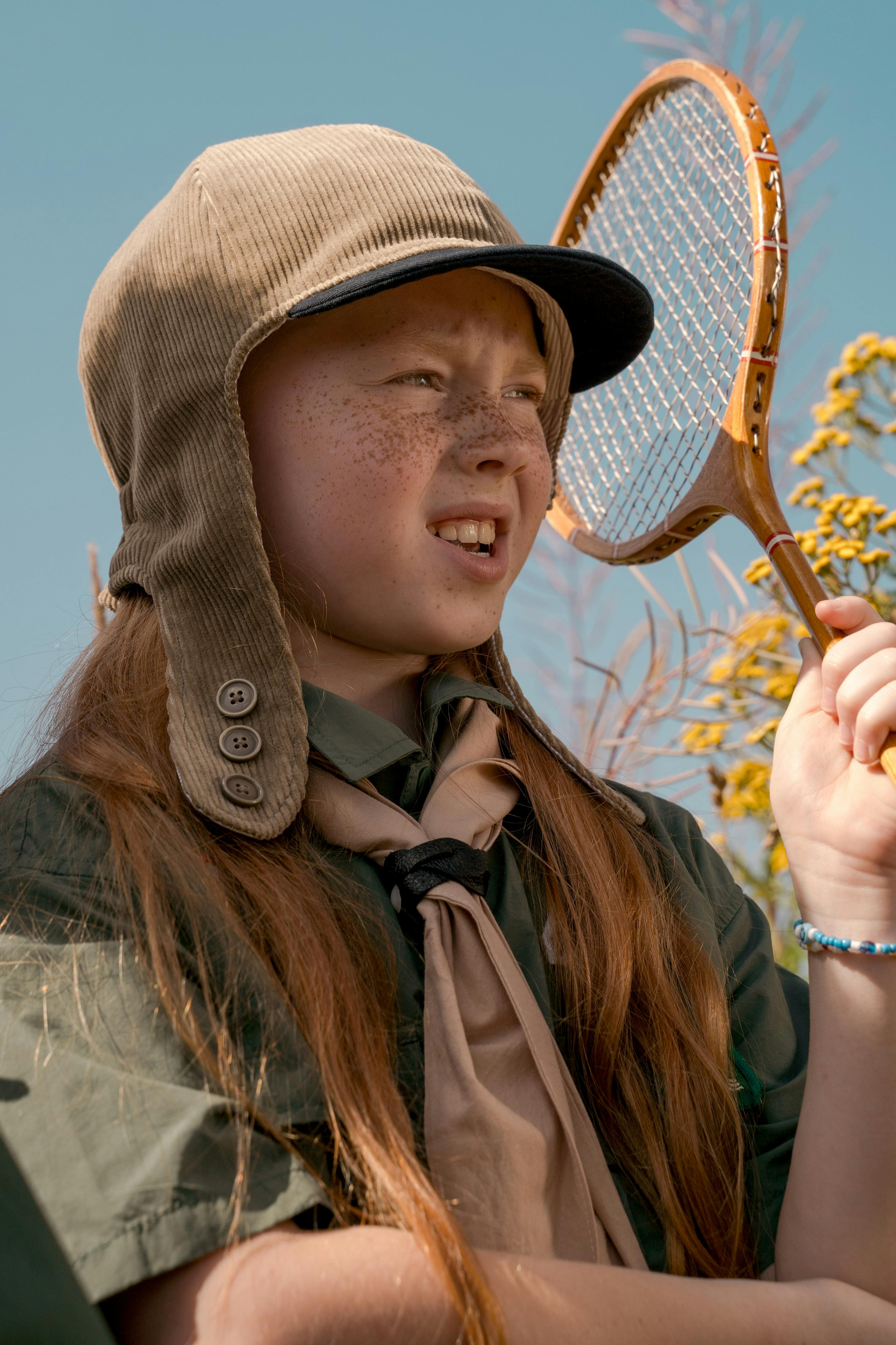 Girl scout wearing uniform and cap poses with a vintage racket on a sunny day.