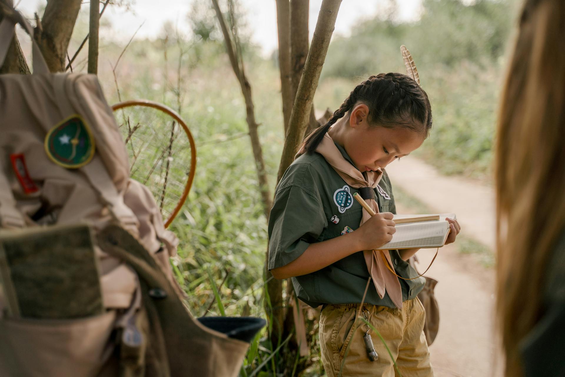 Asian girl scout writing in notebook outdoors, capturing nature notes.