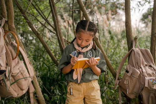 Girl in Grey Shirt and Brown Pants Writing on Yellow Notebook