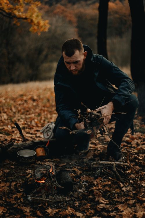 Free A man crouches to build a campfire surrounded by autumn leaves in a forest setting. Stock Photo