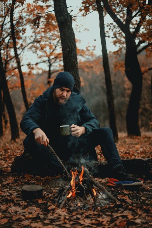 Free Young man enjoys leisure by a campfire in an autumn forest, sipping coffee and relaxing outdoors. Stock Photo