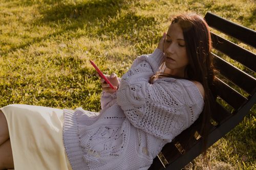Free A woman in a sweater relaxes on a wooden chair outdoors, using her smartphone. Stock Photo