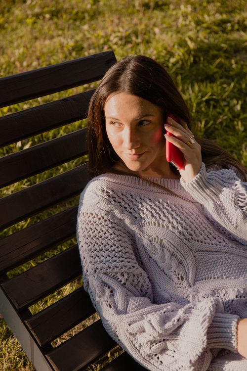 Free Woman in cozy sweater on a sunlit bench, speaking on cellphone outdoors. Stock Photo