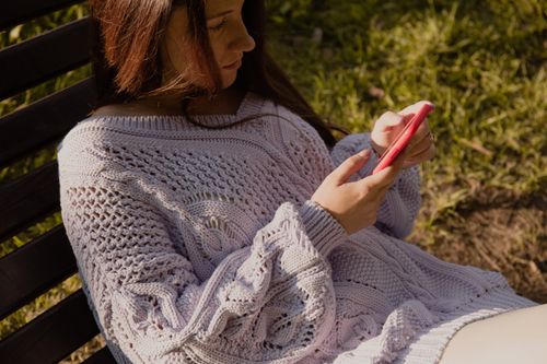 Free Woman in cozy sweater sitting on a wooden bench, using a smartphone outdoors. Stock Photo