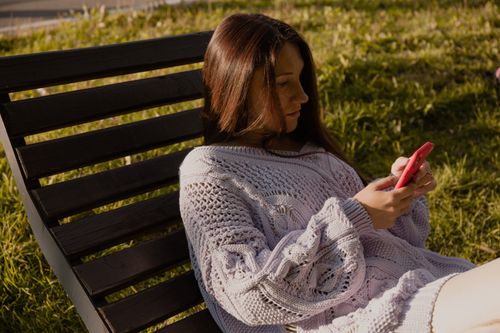 Free A woman in a knitted sweater using a smartphone while relaxing on a wooden chair outdoors. Stock Photo