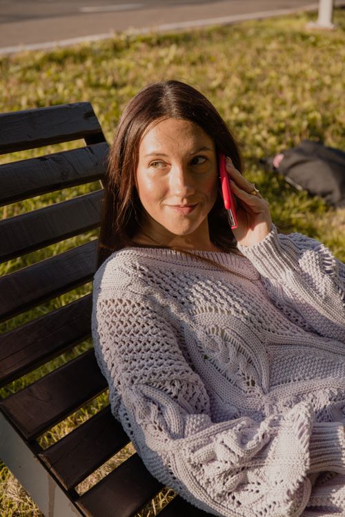 Free Woman in a knitted sweater talking on a smartphone while relaxing outdoors on a wooden bench. Stock Photo