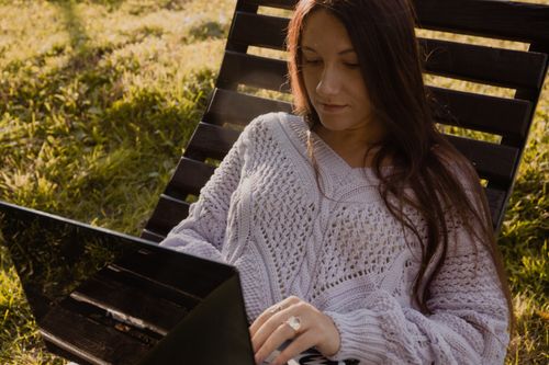 Free Woman in knitwear using laptop outside on a sunny day, enjoying leisure time. Stock Photo