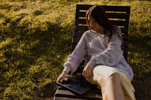Free Young woman in a cozy knitted sweater using a laptop while relaxing outdoors in sunlight. Stock Photo