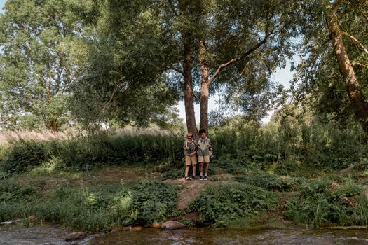 Two children in scout uniforms stand together by a forest stream, enjoying outdoor exploration.