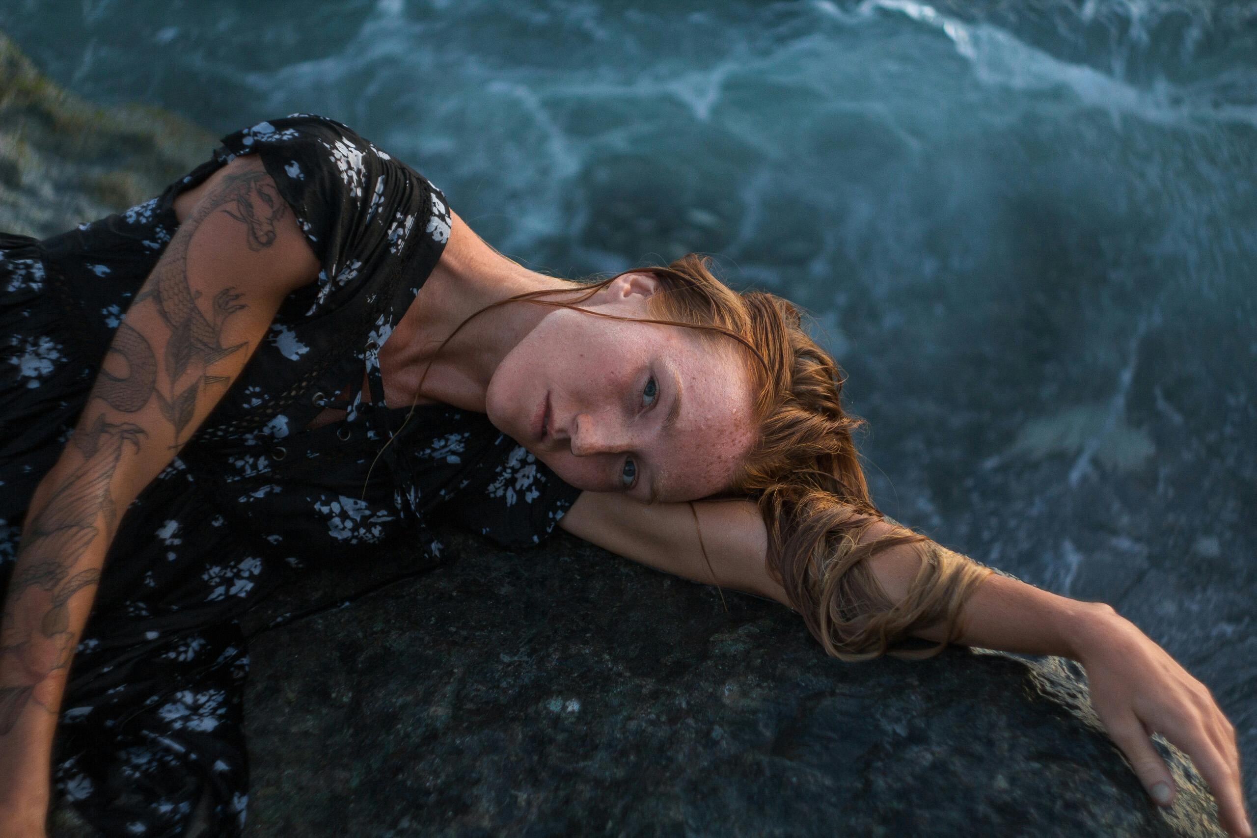 Caucasian woman with tattoos relaxing on a rock by the seaside in a dreamy pose.