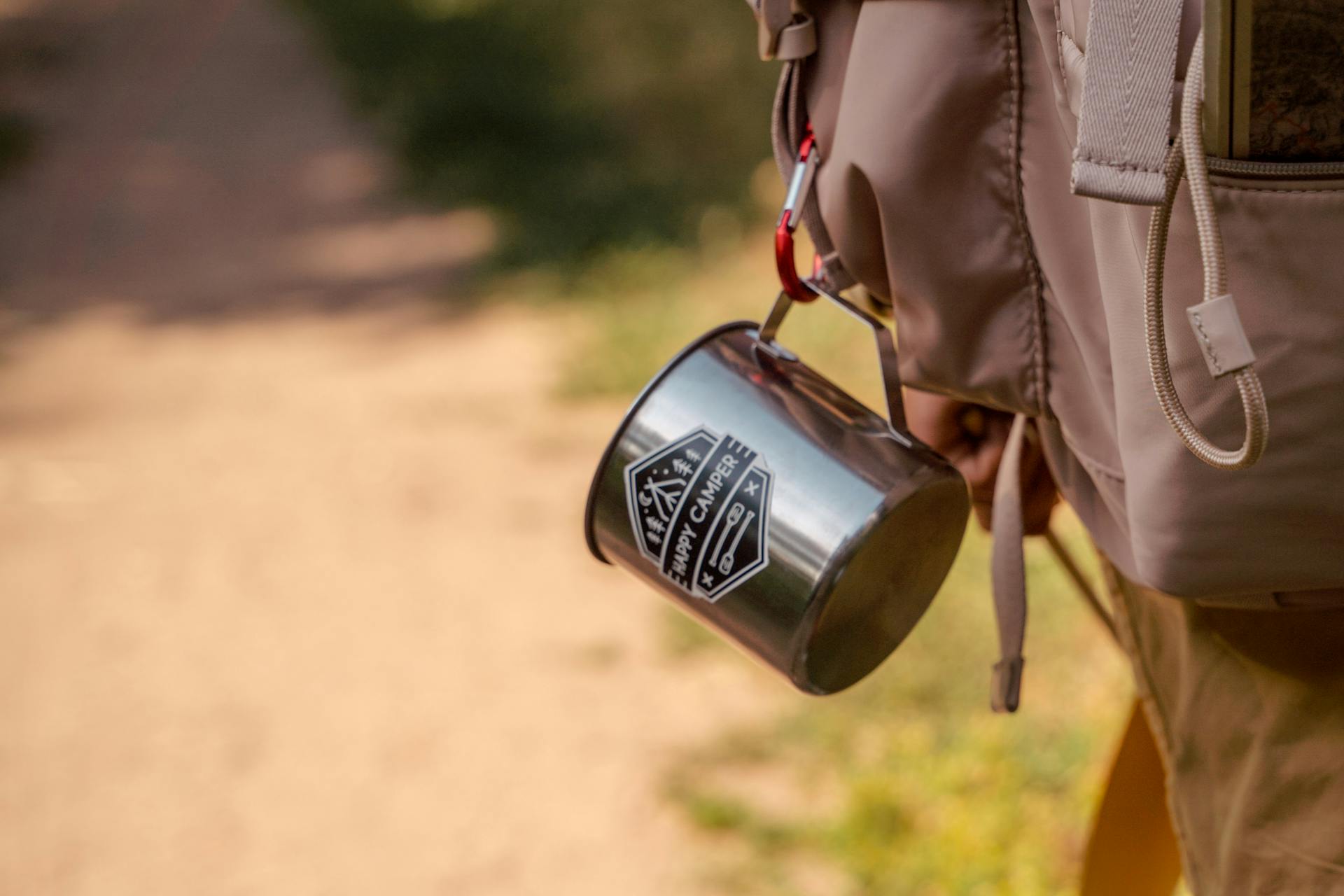 Stainless steel camping mug clipped to backpack with carabiner on hiking trail.