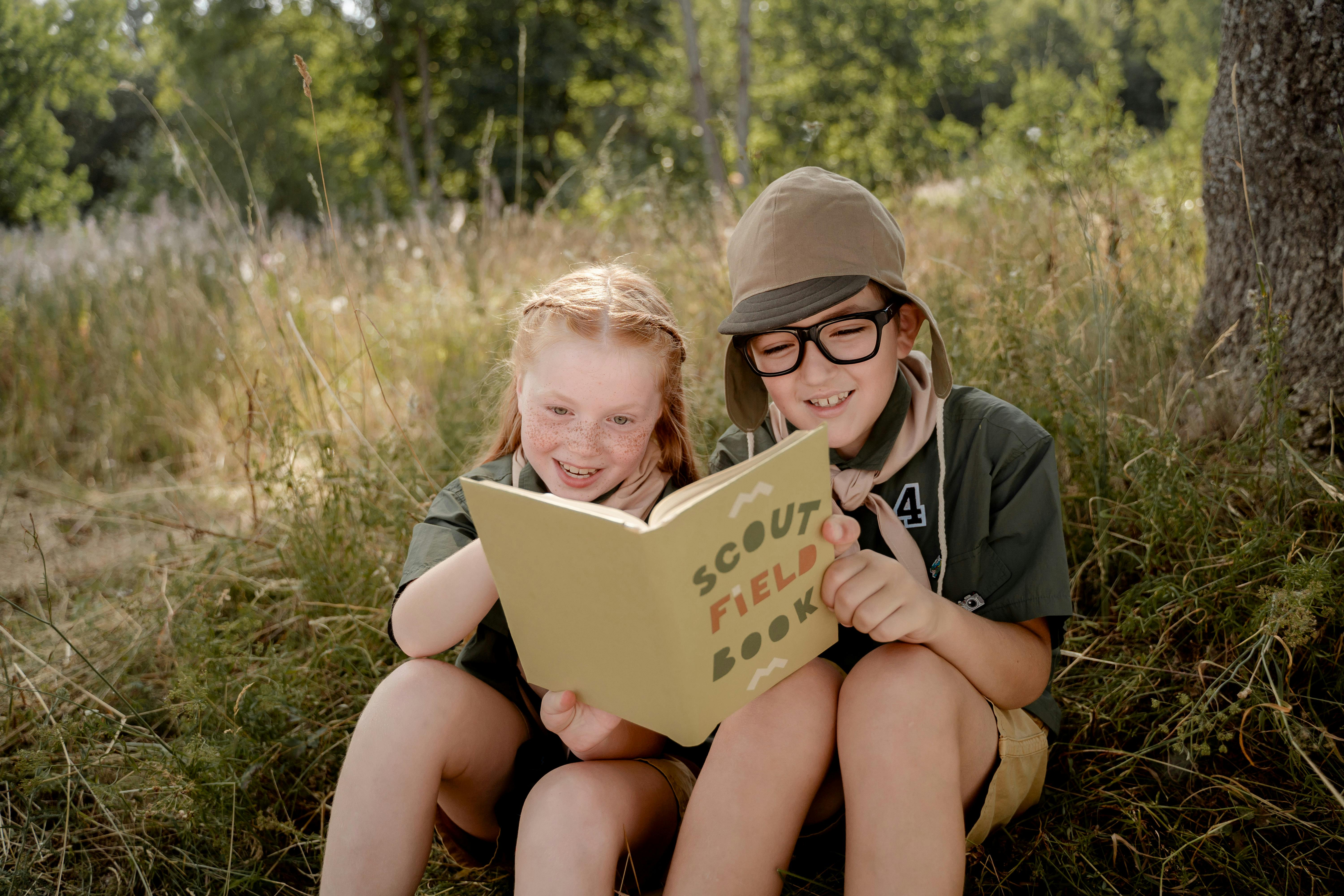 Two children in scout uniforms reading a field book in a forest setting.