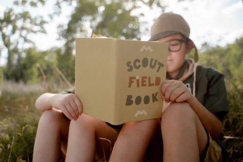 Free A young scout wearing glasses sits in a field reading a field book on a sunny day. Stock Photo