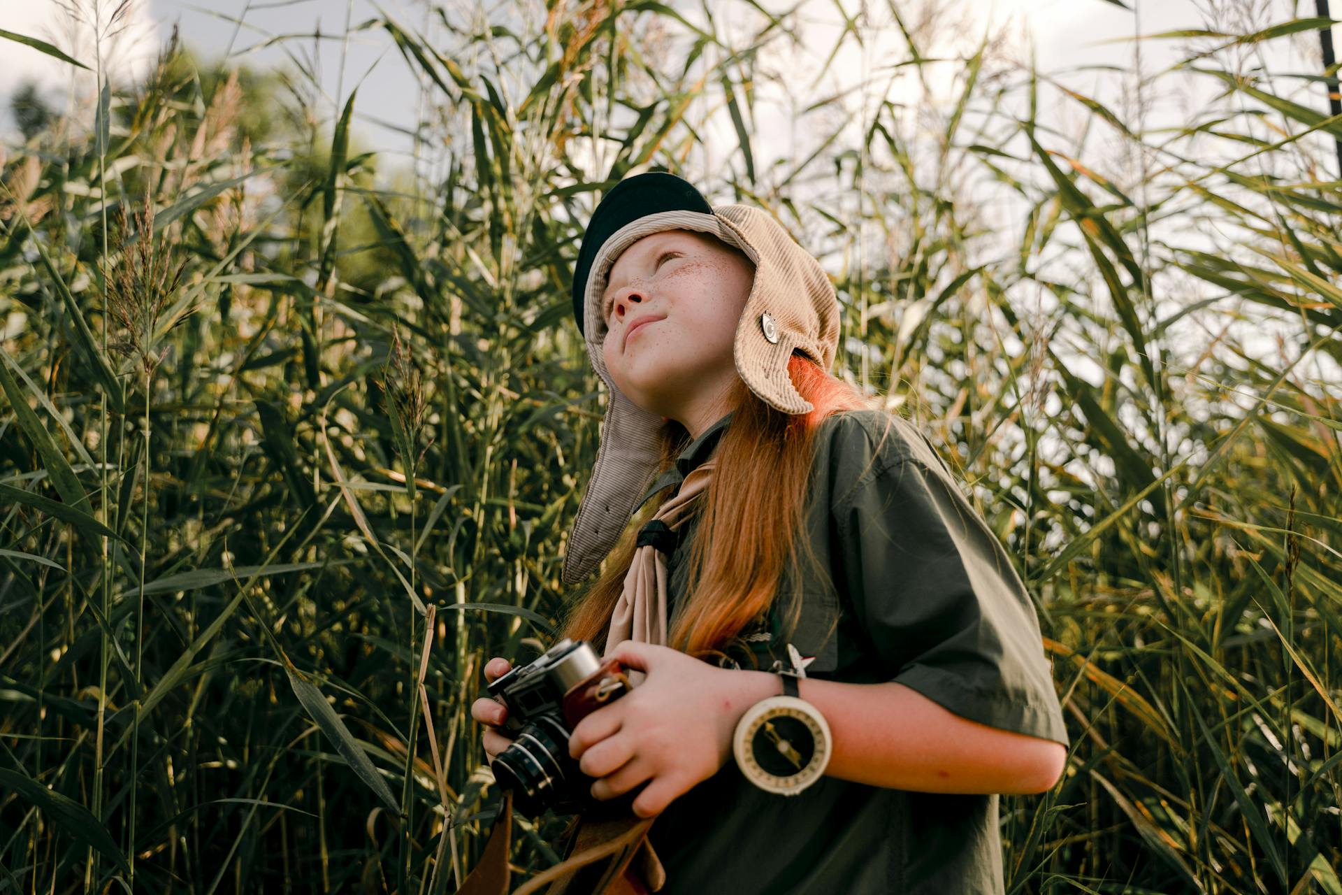 Young girl scout with camera in nature, looking up amidst tall grass.