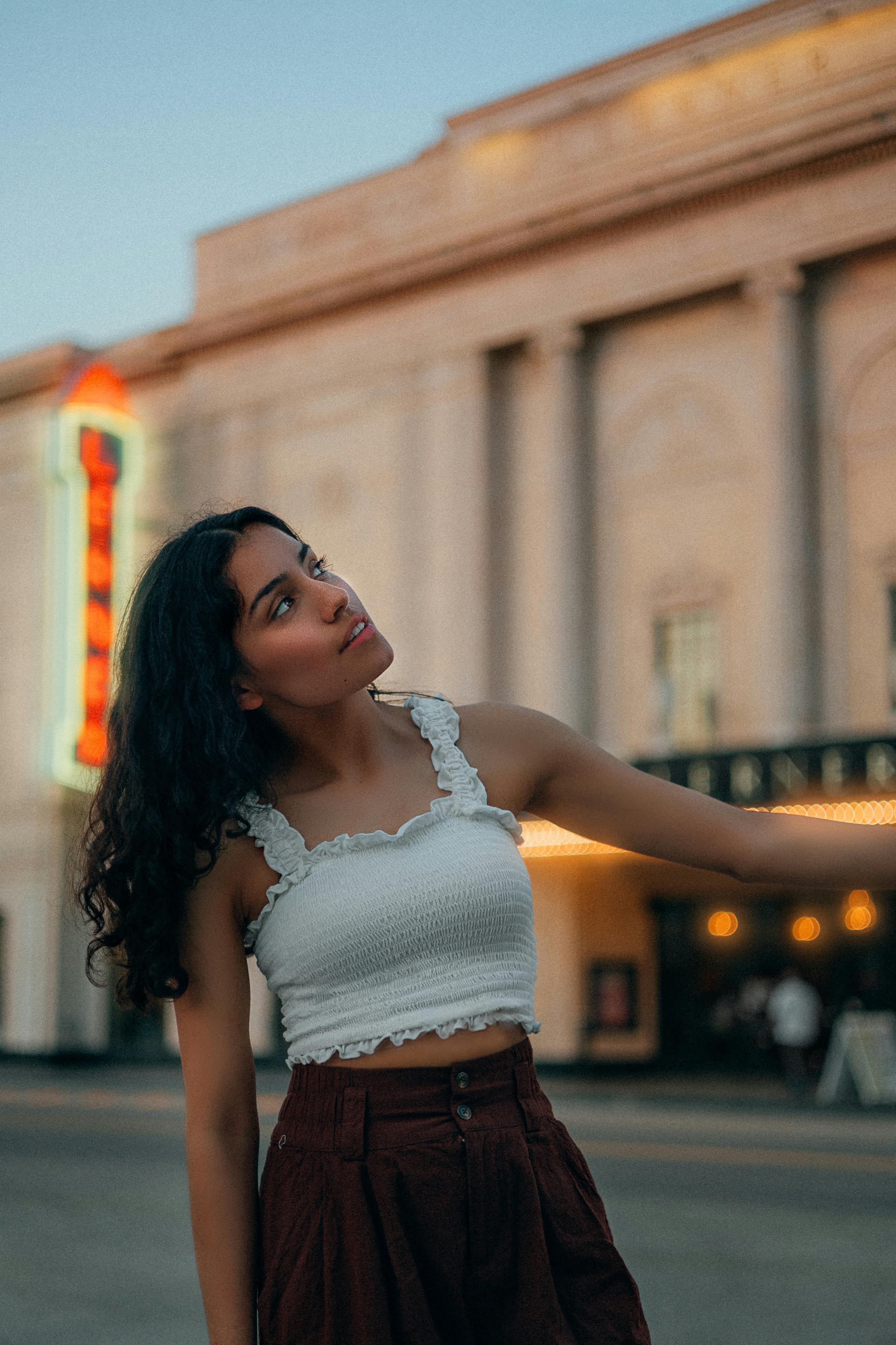 Free A young woman in stylish clothing poses thoughtfully in front of a historic theater during twilight. Stock Photo