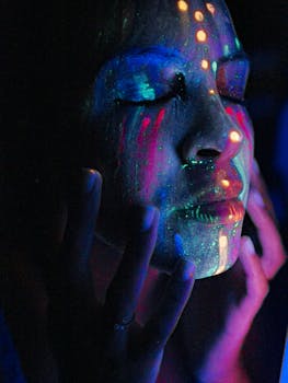 A woman with colorful neon face paint under blacklight, eyes closed, in the dark.