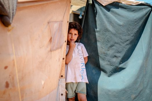 A young child stands by a shelter in a Syrian refugee camp, looking thoughtful.