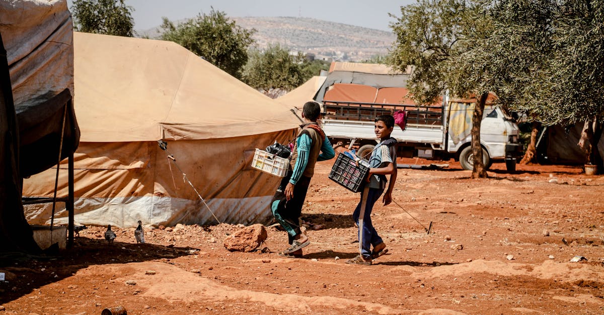 Two boys carrying crates walk through a refugee camp in Idlib, Syria, amidst tents and trees.
