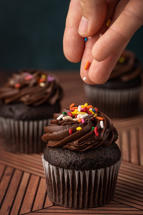 
A Close-Up Shot of a Person Putting Sprinkles on a Chocolate Cupcake