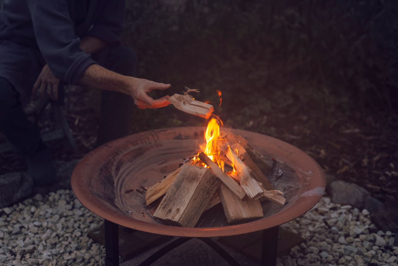 Person adding firewood to a round steel fire bowl with flames