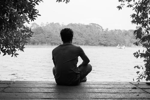 Gray Scale Photography of Man Sitting on Brown Wooden Floor Beside Body of Water