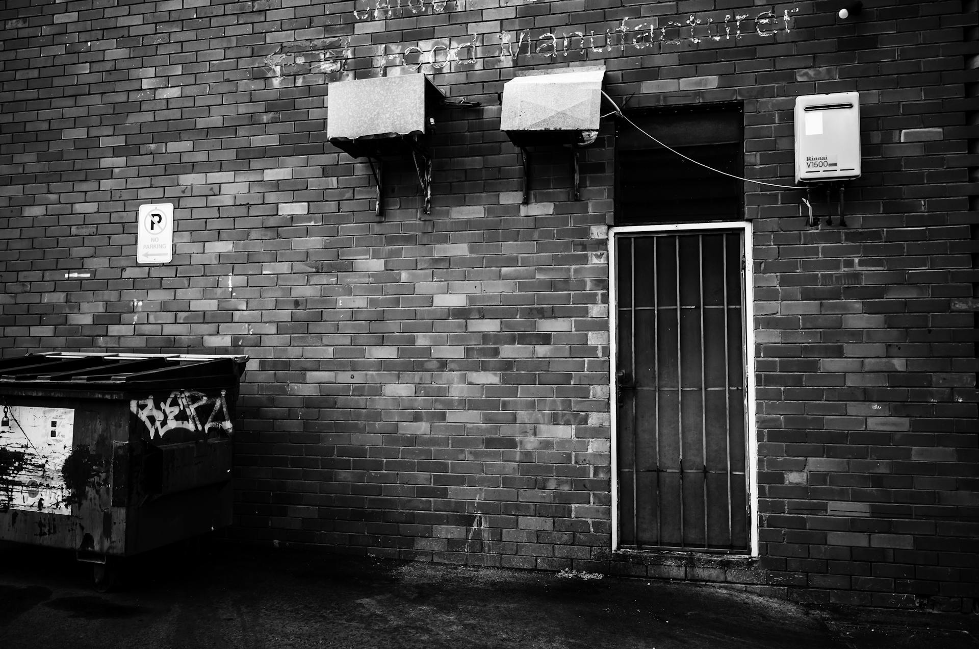 A gritty black and white photo of a back entrance with a brick facade and graffiti-covered trash can.