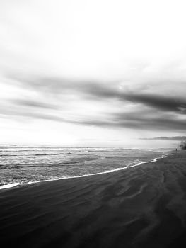 Black-and-white beach scene with dramatic clouds and gentle waves on the horizon.