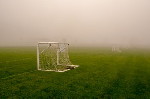 Misty view of an empty soccer field with visible goalposts, surrounded by thick fog.