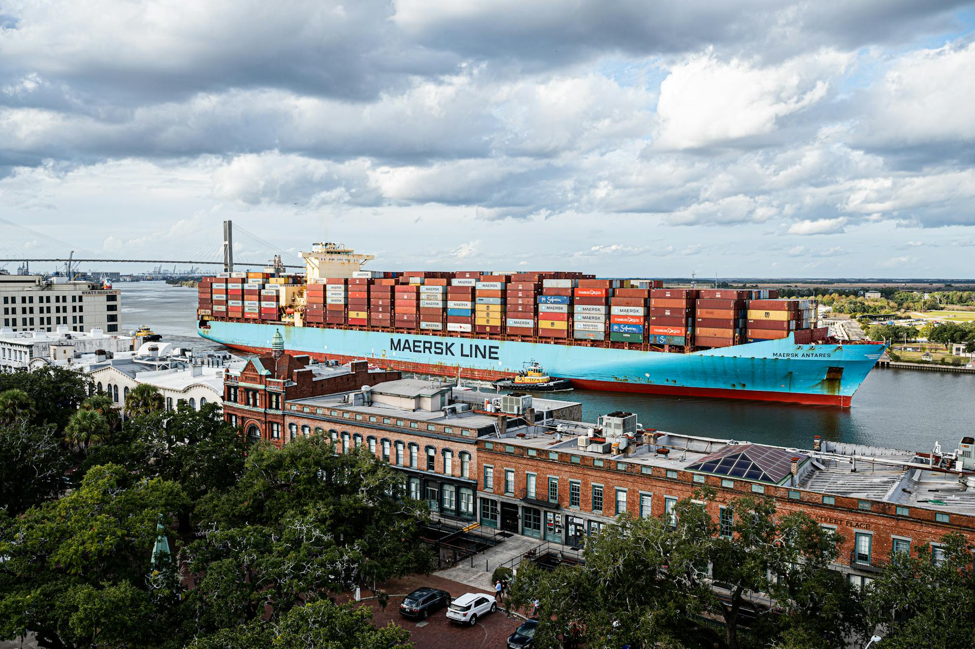 Aerial view of a cargo ship on the Savannah River with a city backdrop and cloudy sky.