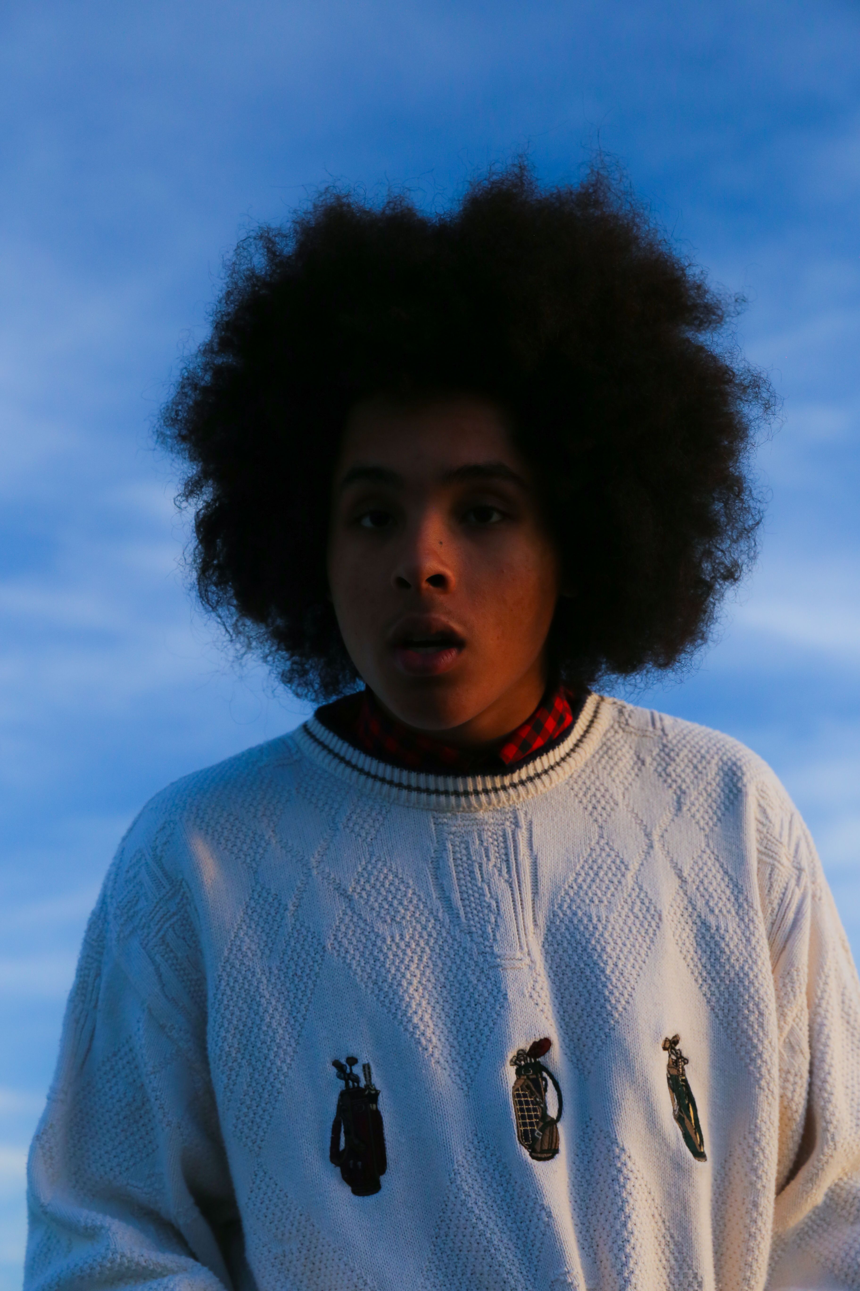 A contemplative portrait of a person with afro hair wearing a white sweater against a clear blue sky.