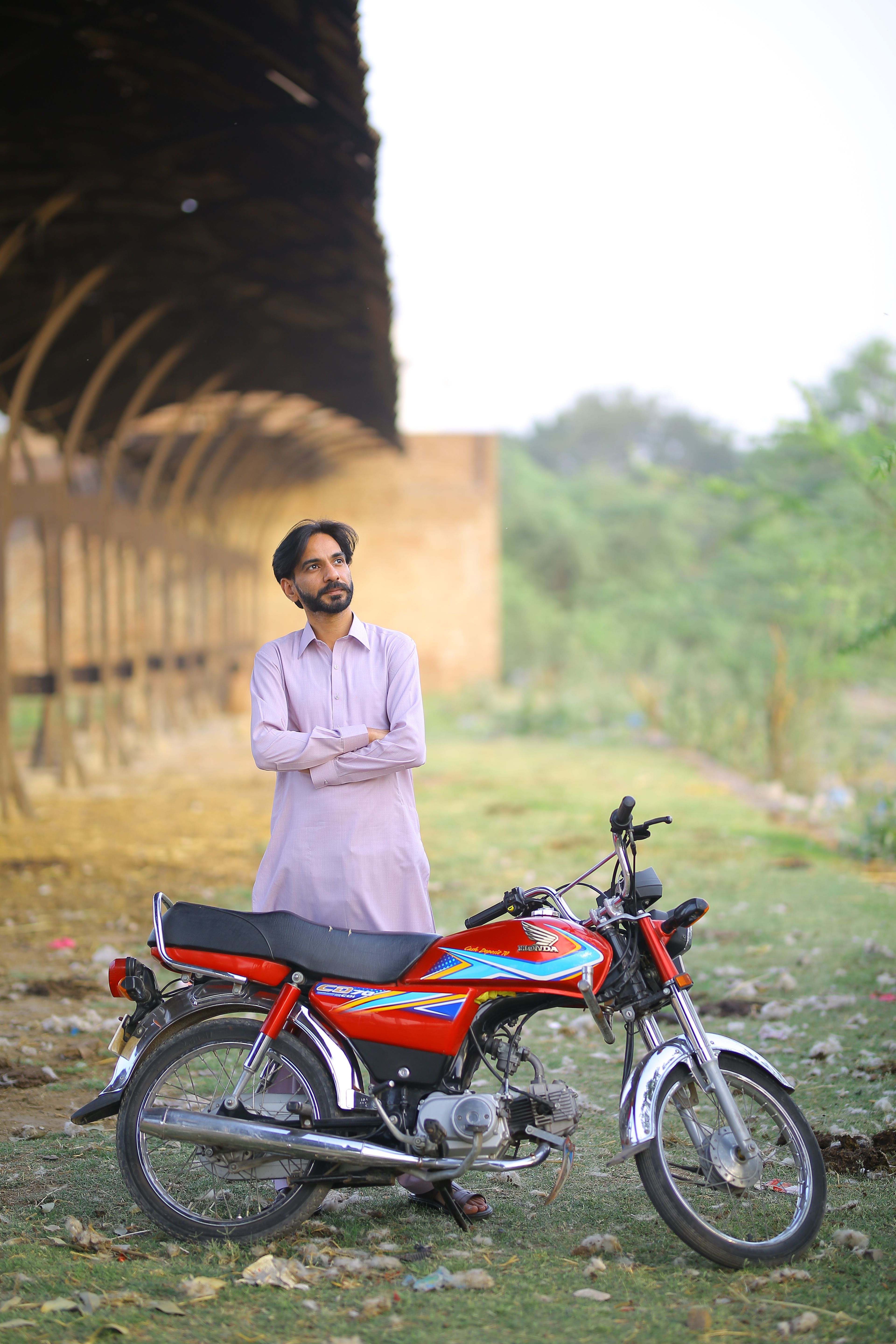 A man in traditional attire stands next to a motorcycle outdoors in Kasur.