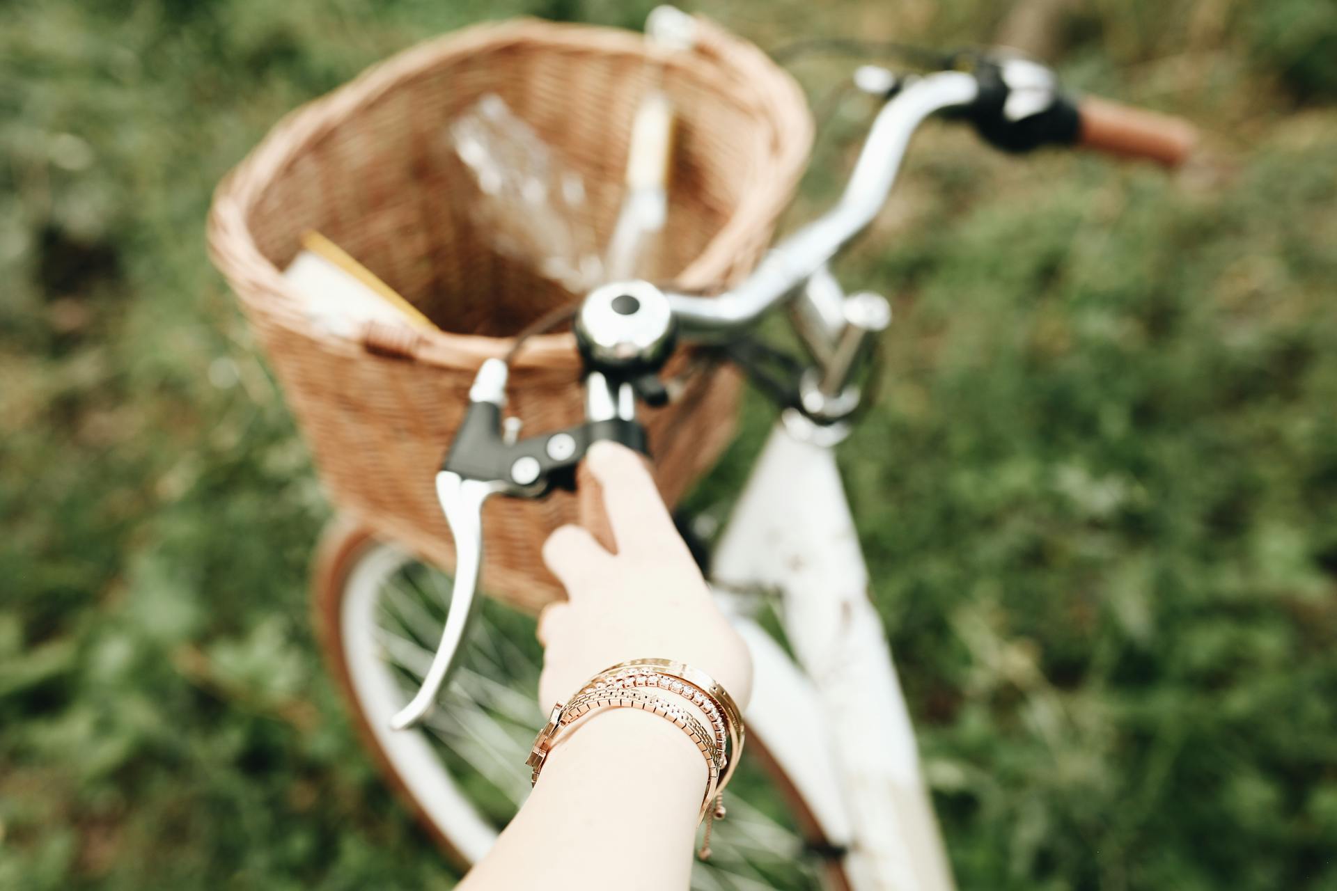A hand grips the handlebar of a bike with a wicker basket in a grassy field.