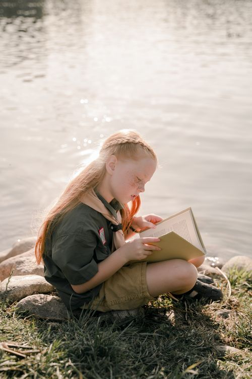 Free A young girl scout reads a book by the tranquil edge of a lake, enjoying a sunny day outdoors. Stock Photo