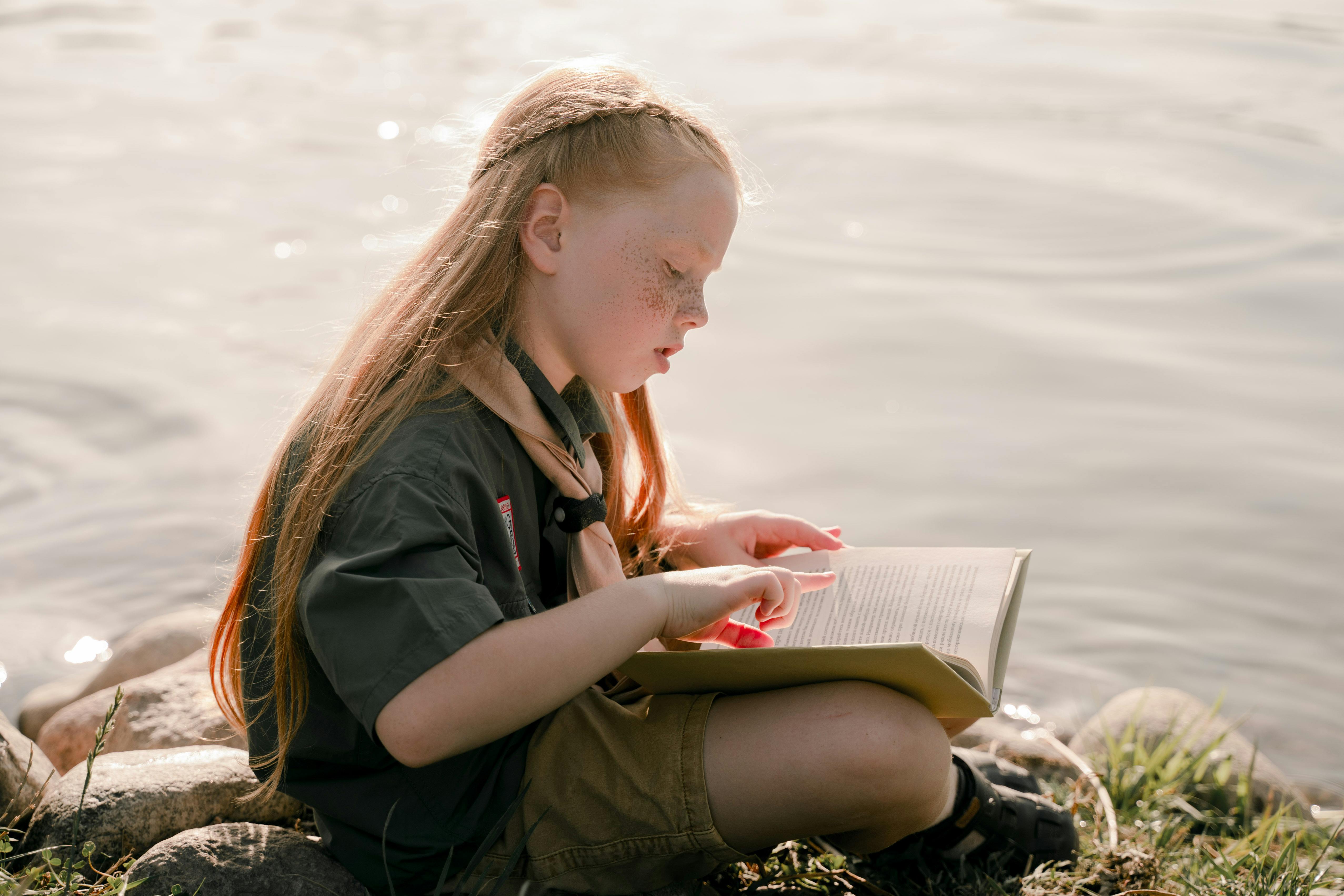 A young girl peacefully reading a book by a calm lakeside, enjoying a sunny day outdoors.