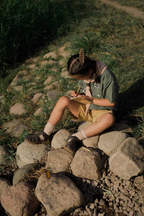 Free Girl in Green Shirt and Brown Shorts Sitting on Rock Stock Photo