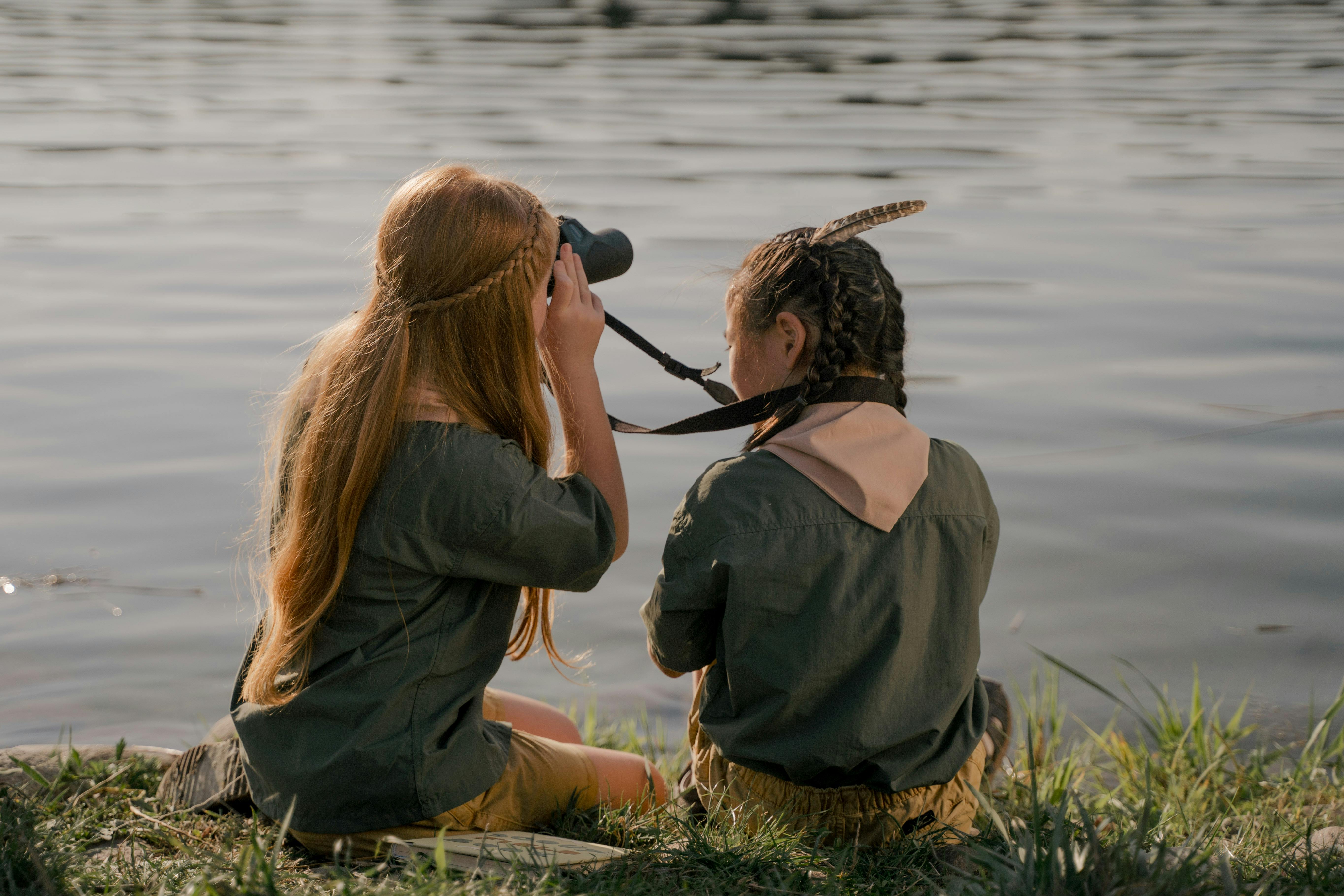 Two young girl scouts sit by a lake, observing birds with binoculars, a serene moment in nature.