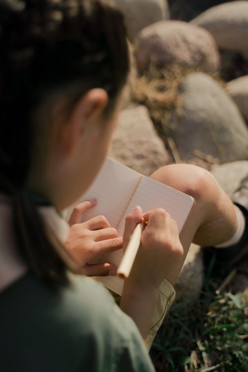 Free A girl in scout uniform writes in a notebook while sitting on rocks outdoors. Stock Photo