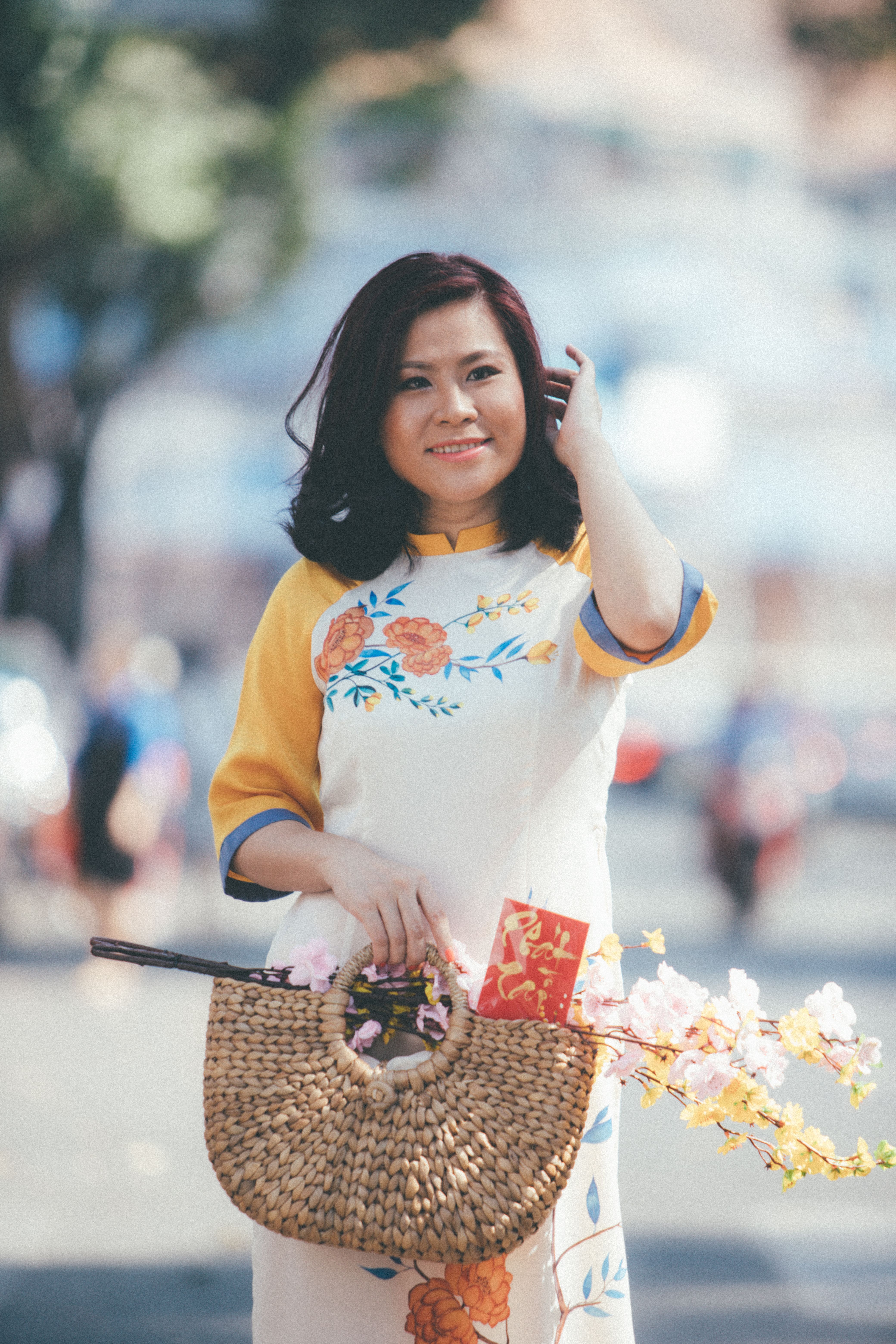 Asian woman in a traditional floral dress smiling and holding a woven bag outdoors. - Budget-Friendly Handbag Options in Pakistan
