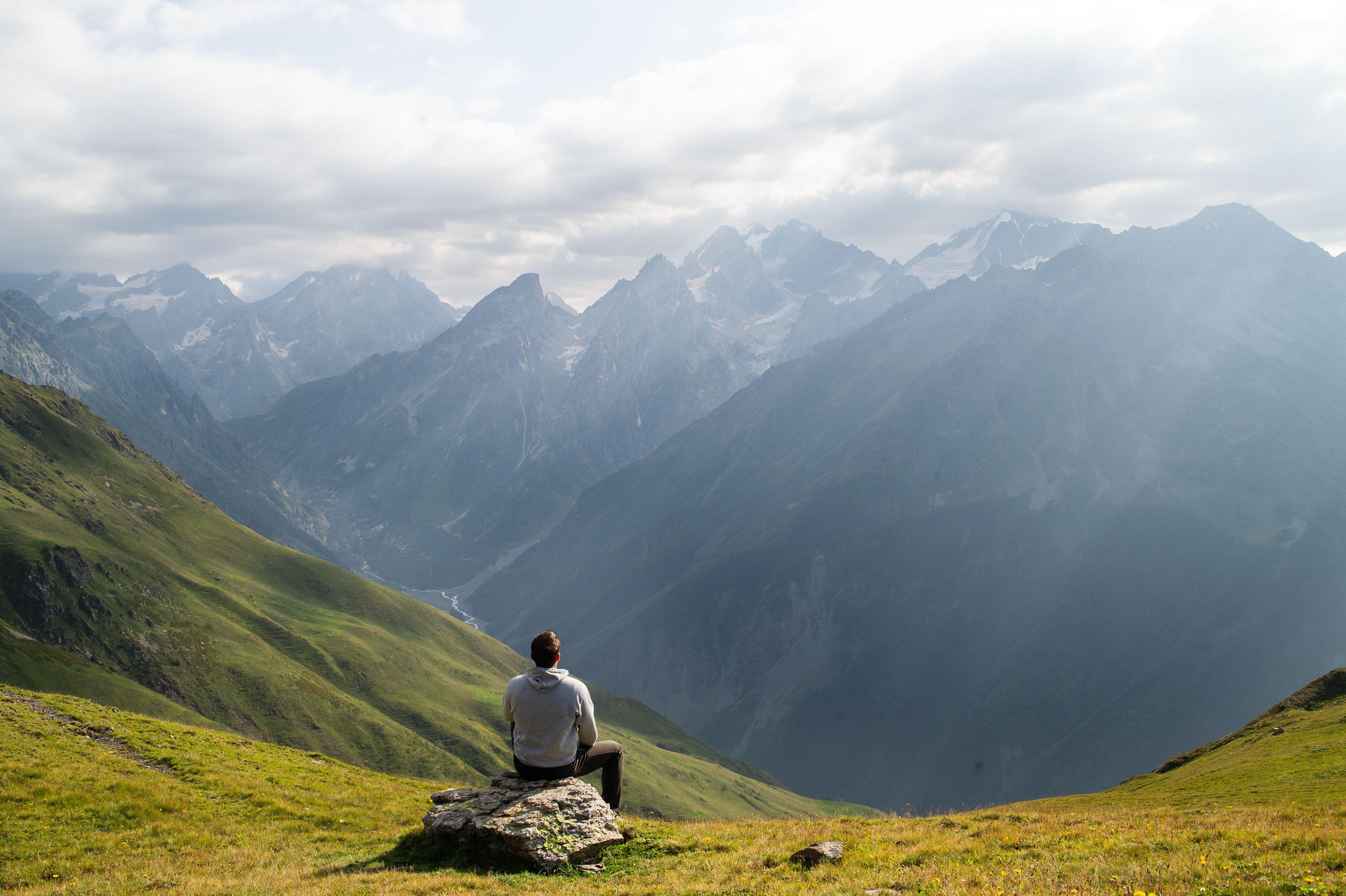 A person sitting on a rock, admiring majestic mountains under cloudy skies.
